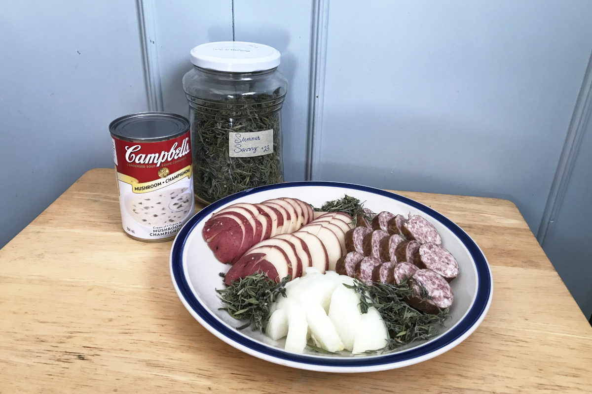 green bean glory ingredients including a can of Campbell's mushroom soup, and a plate of sliced potatoes, diced onion and sliced farmer sausage garnished with summer savory