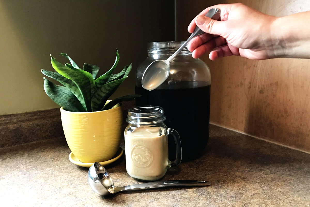 Mug of cold brew coffee, milk, and spoon dripping with sweetened condensed milk, and large jar of cold brew coffee and potted plant.