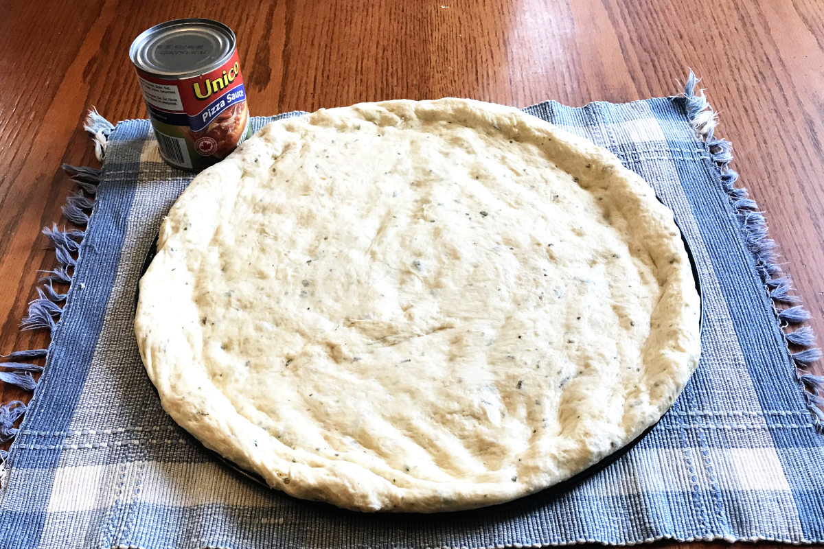 homemade pizza dough spread out on pizza pan on placemat with can of pizza sauce on the side