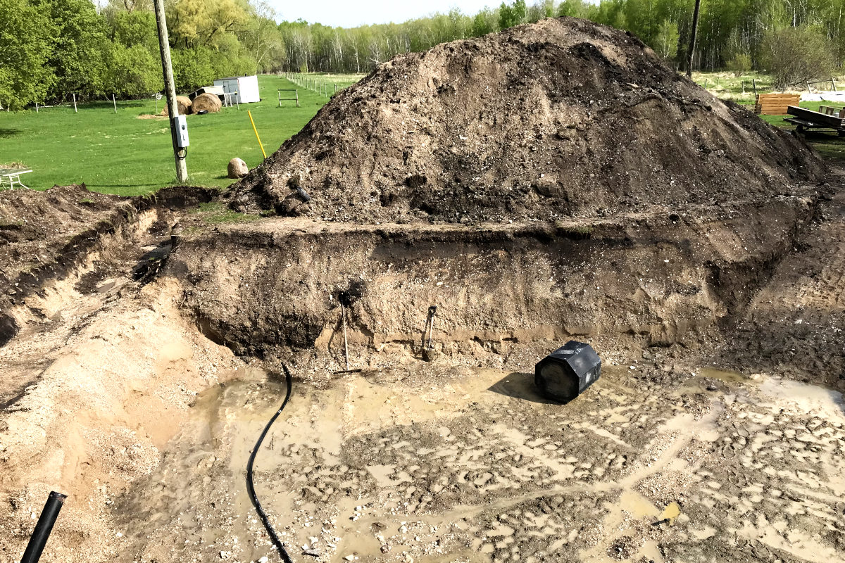 excavated hole for our old house remodel with most of the water drained, and a large pile of gravel and dirt behind it.