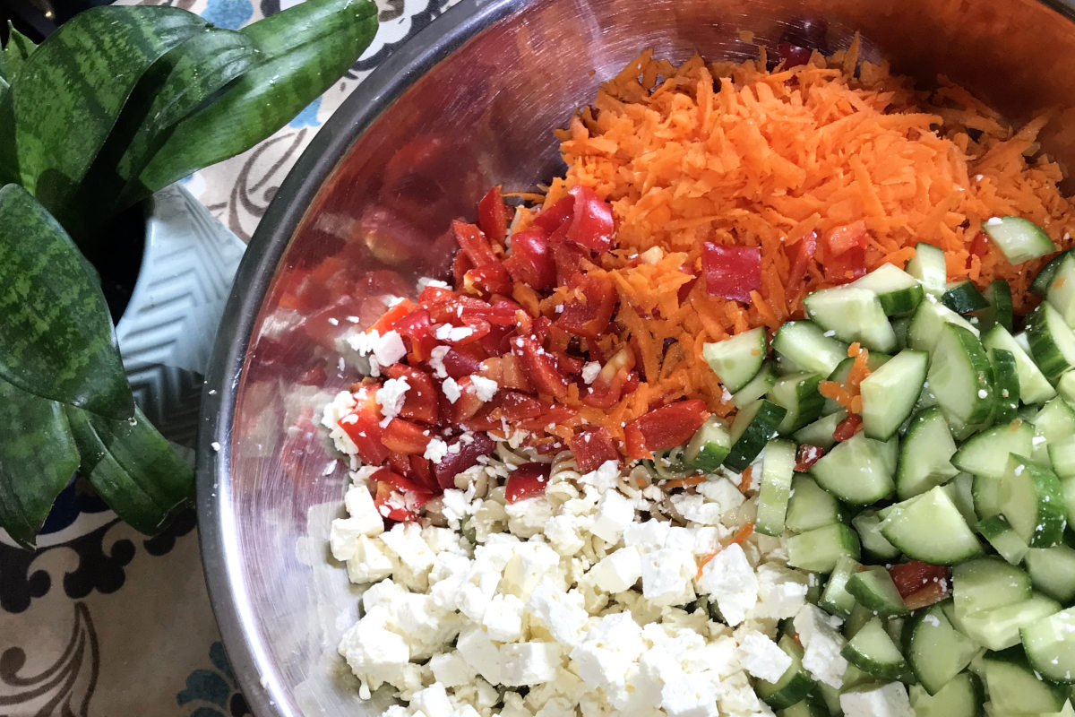 stainless steel bowl with chopped ingredients for ultimate macaroni salad, including chopped red pepper, grated carrots, chopped cucumber and diced feta