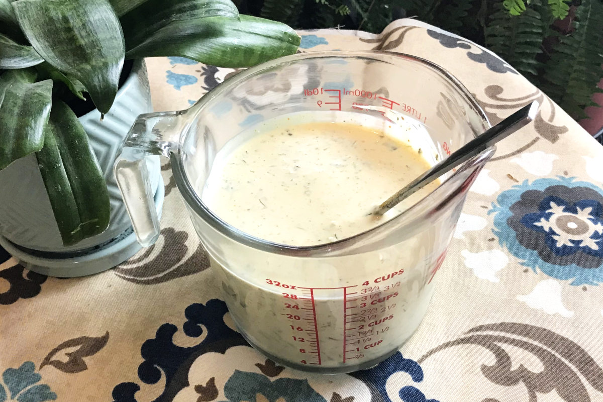 measuring cup of dressing for the ultimate macaroni salad on table cloth with a plant