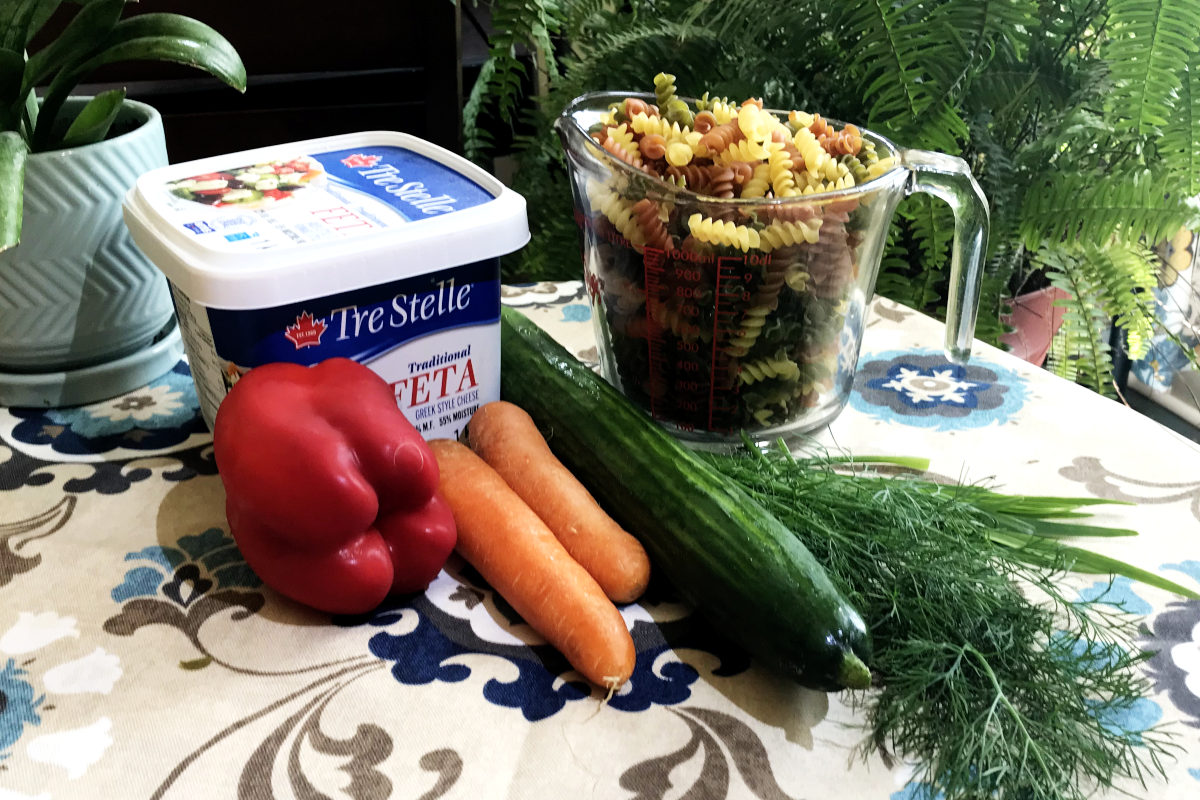 ingredients for making the ultimate macaroni salad, including feta in a container, a red pepper, two carrots, an English cucumber, a bunch of dill and garlic chives, and a measuring cup of dry macaroni, all on a table cloth with plants in the background