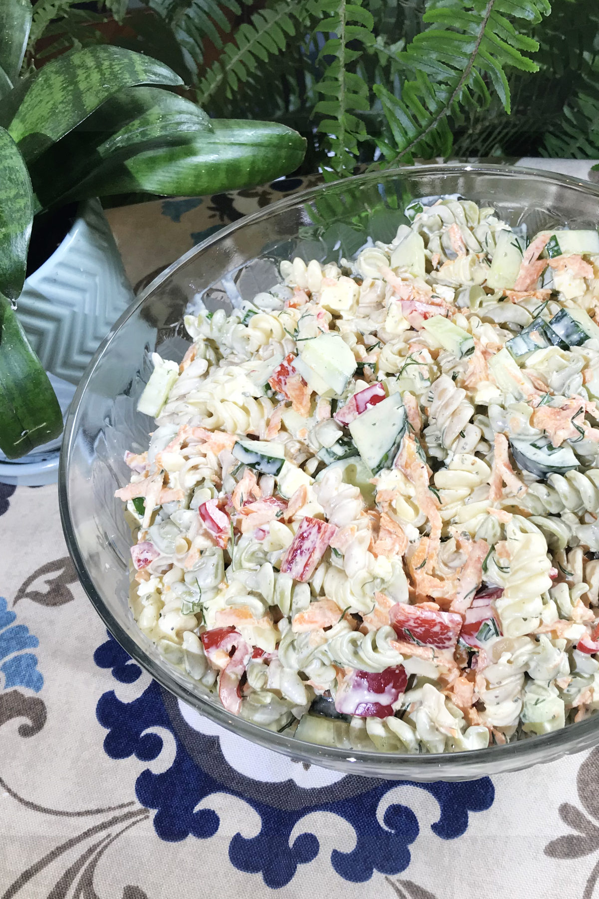 Ultimate Macaroni Salad in a glass serving bowl with plants in the background
