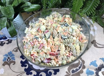 glass serving bowl of the ultimate macaroni salad on a table cloth with plants in the background