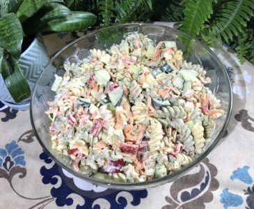 glass serving bowl of the ultimate macaroni salad on a table cloth with plants in the background