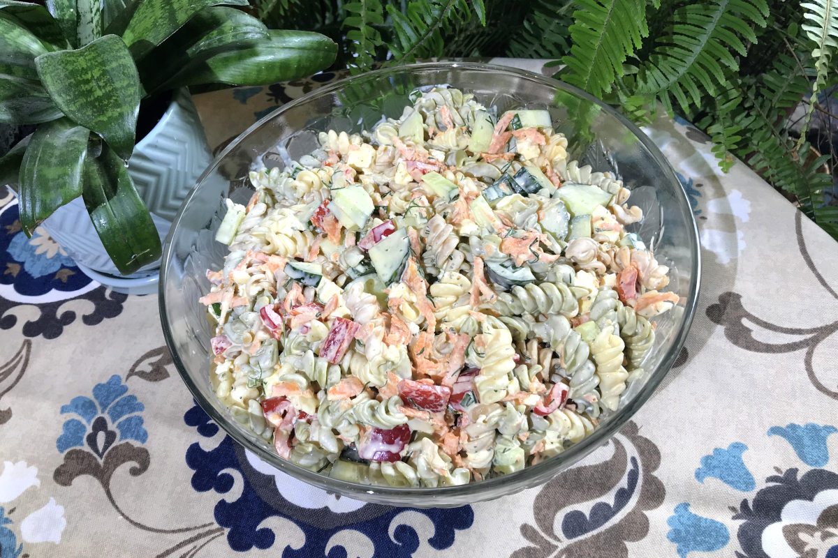 glass serving bowl of the ultimate macaroni salad on a table cloth with plants in the background