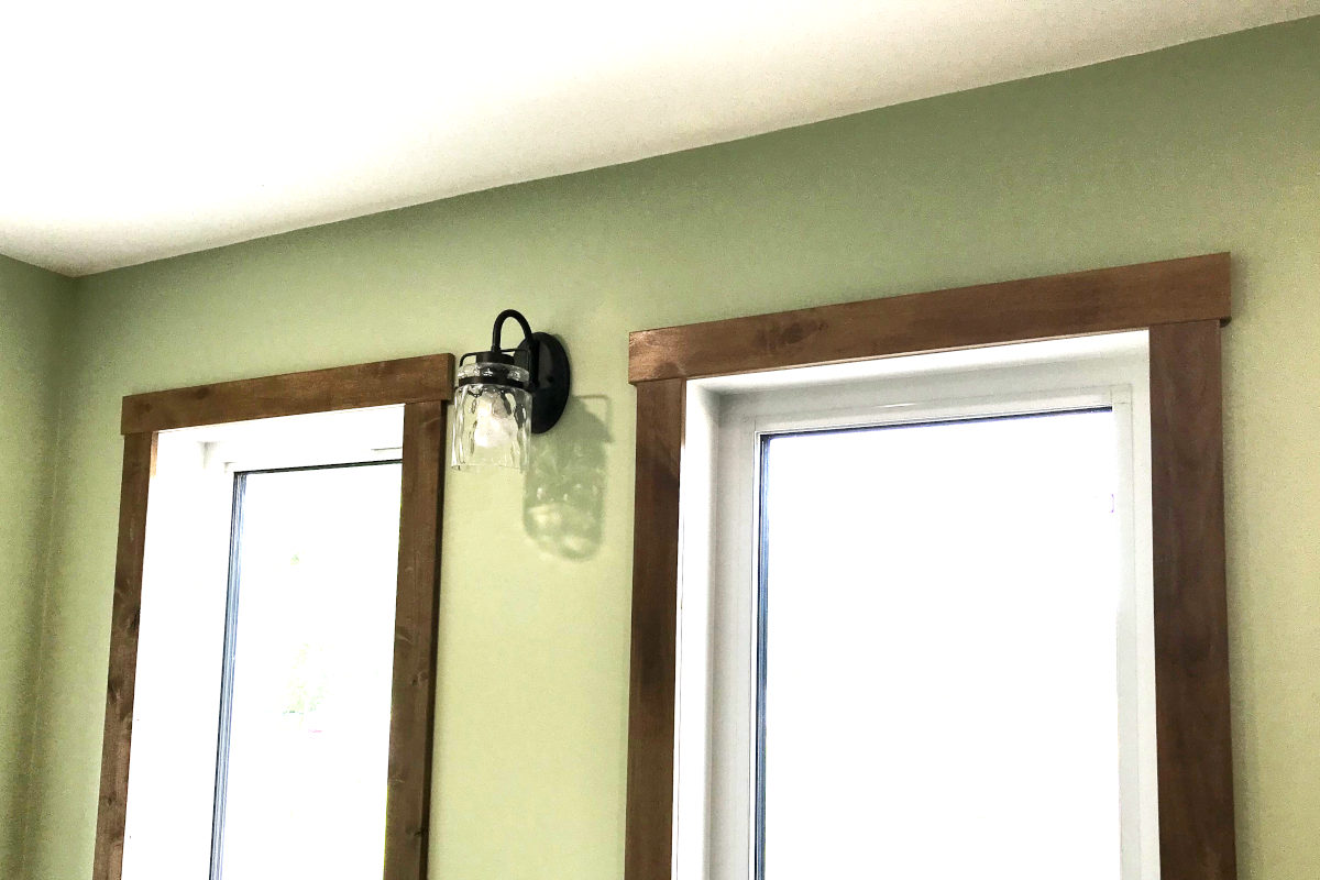 wood stained casing around tall windows with sage green walls and a black sconce with clear glass