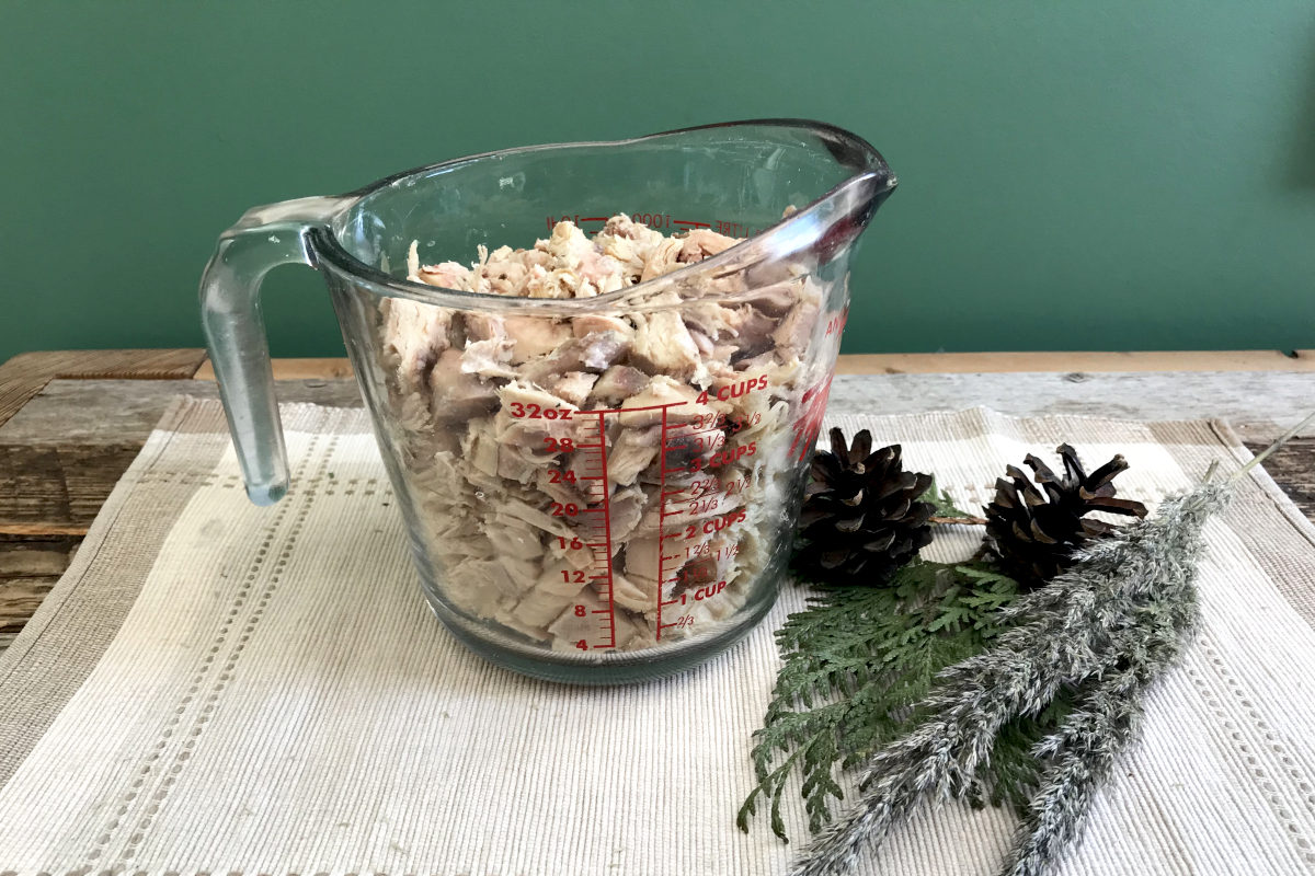 Four cup measuring cup of chopped chicken on a place mat with greenery on weathered wood