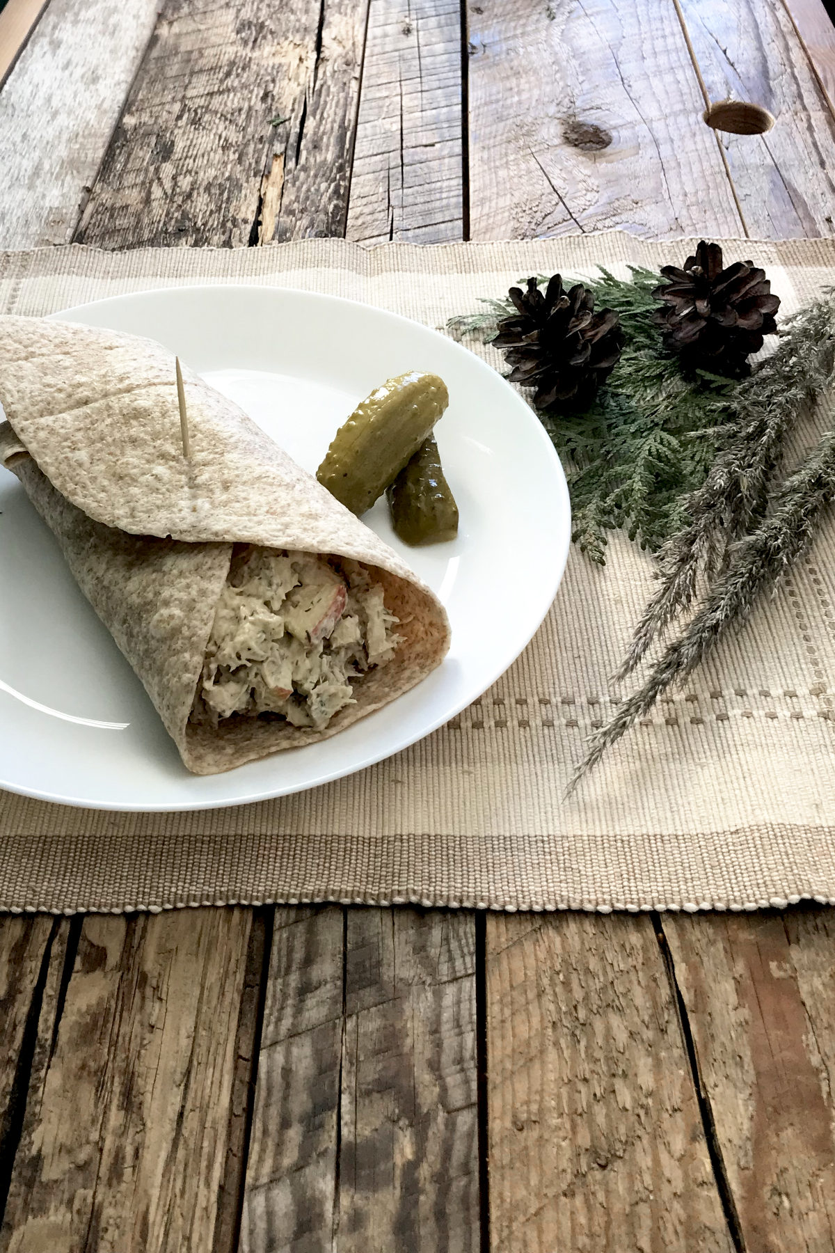 Homemade Chicken Salad in a torilla wrap on a white plate with pickles, on a place mat with greenery, on weathered wood