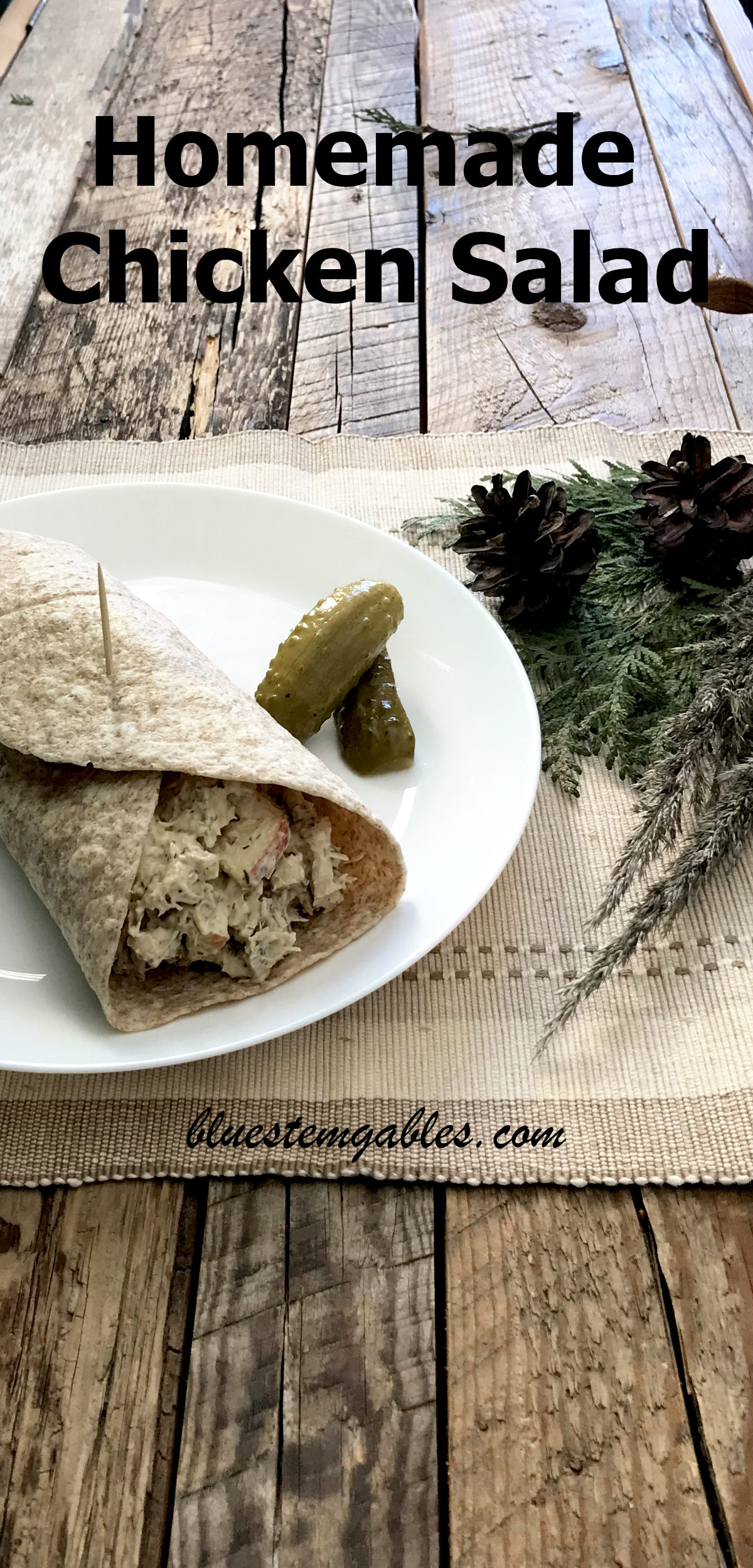Homemade chicken salad tortilla wrap on a white plate with pickles, on a place mat with greenery on weathered wood with text reading homemade chicken salad