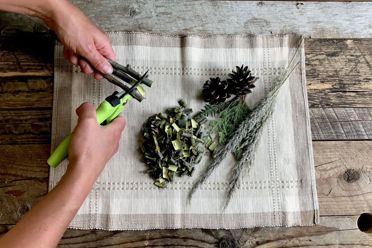 using pruning shears to cut the fresh willow bark into small pieces, on a place mat with greenery on weathered wood