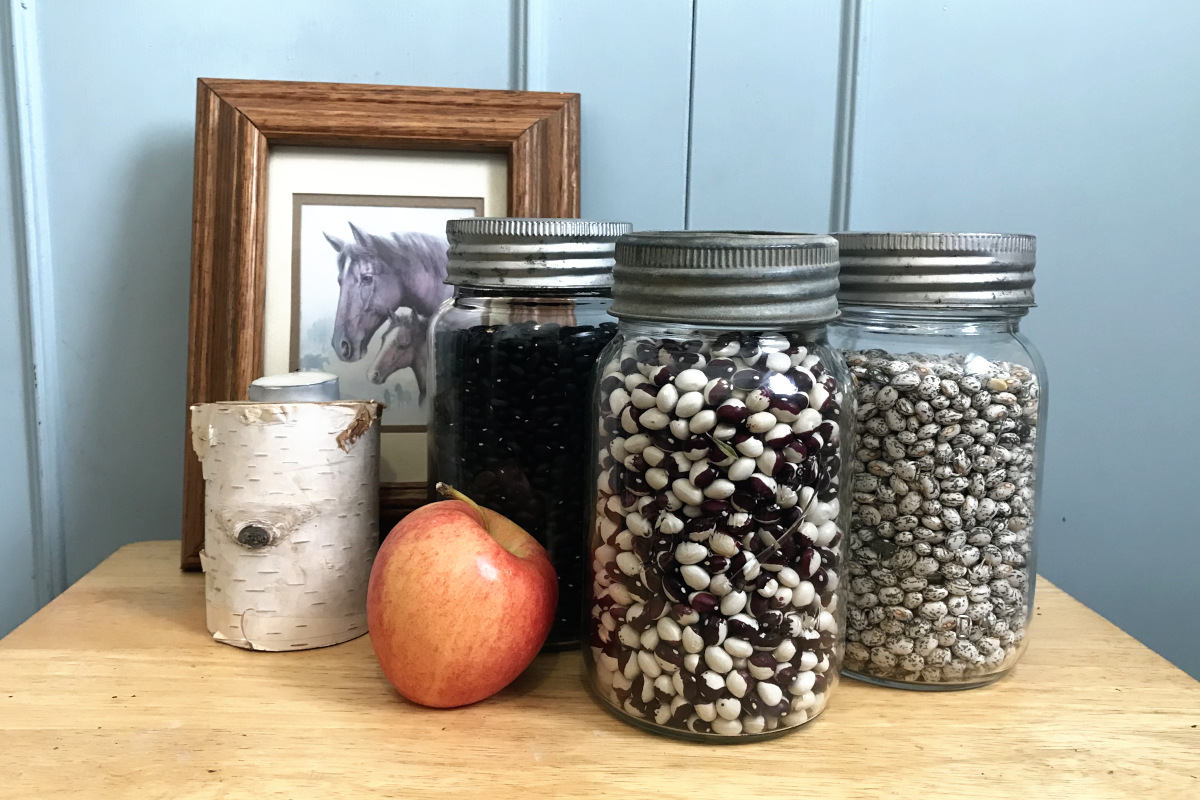 three jars of different dry beans with horse picture, birch candle, and apple on a wood table