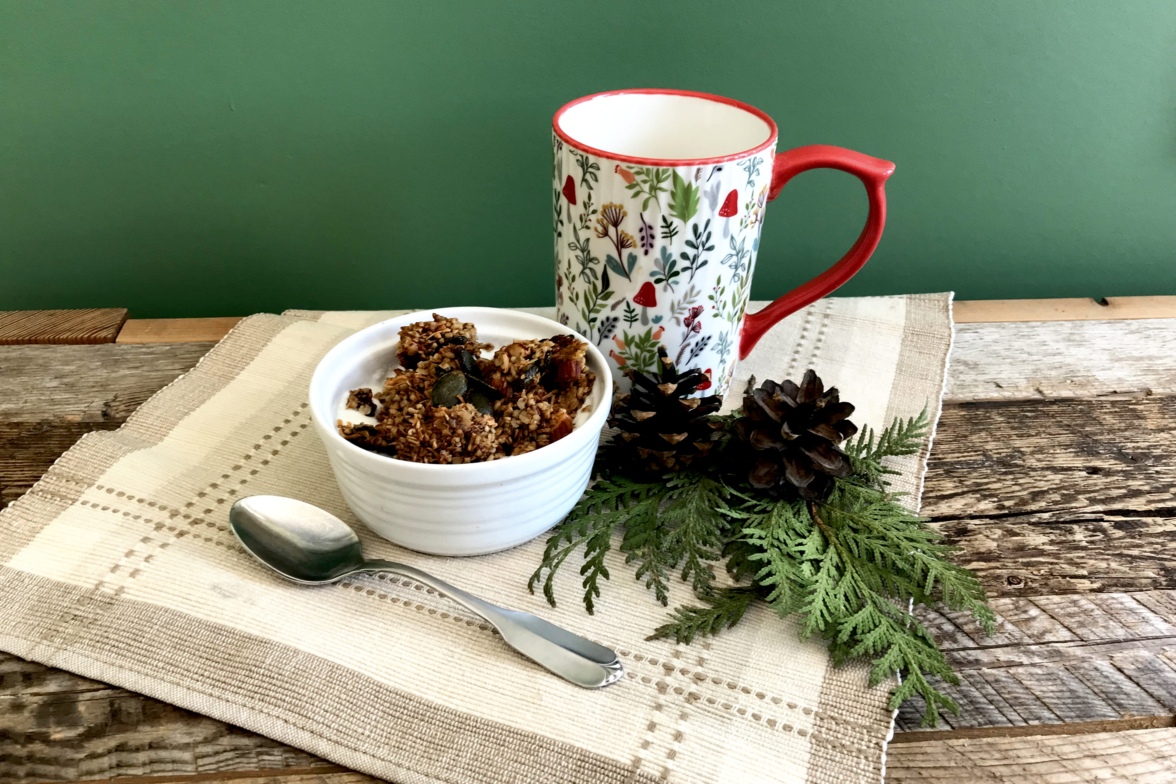 bowl of homemade granola on a place mat with a mug and greenery on weathered wood