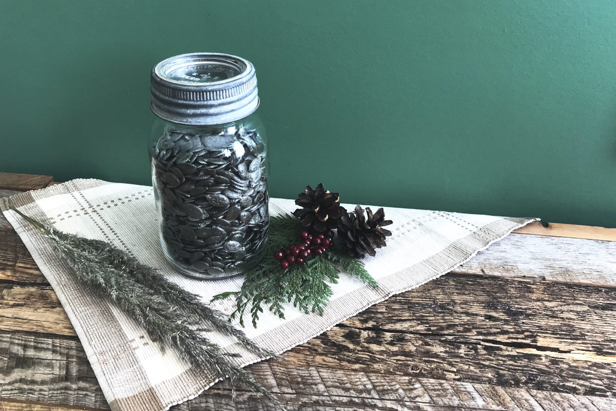 jar of hulless pumpkin seeds on placemat on weathered wood with greenery