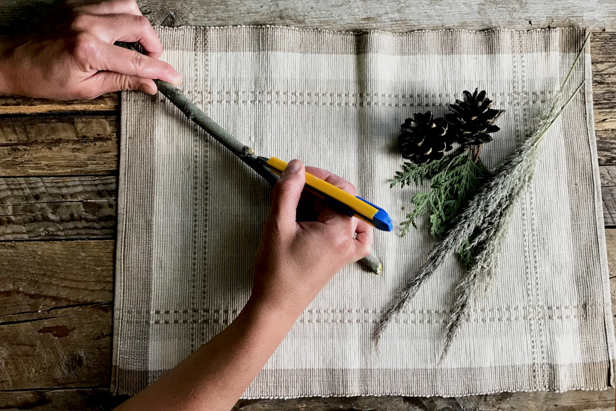 slicing willow bark from a small branch with a utility blade, on a place mat with greenery on weathered wood