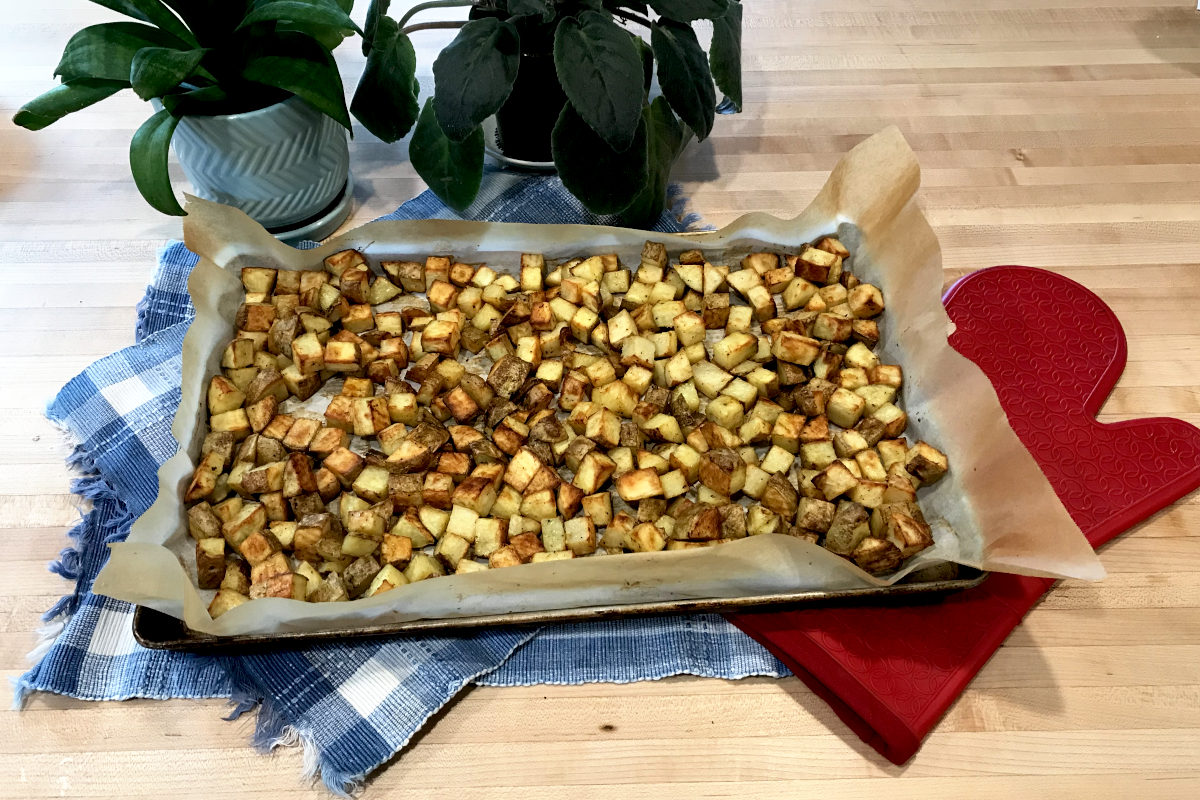 Baked, diced potatoes on baking sheet with parchment paper on place mats with oven mitt and potted plants on butcher block