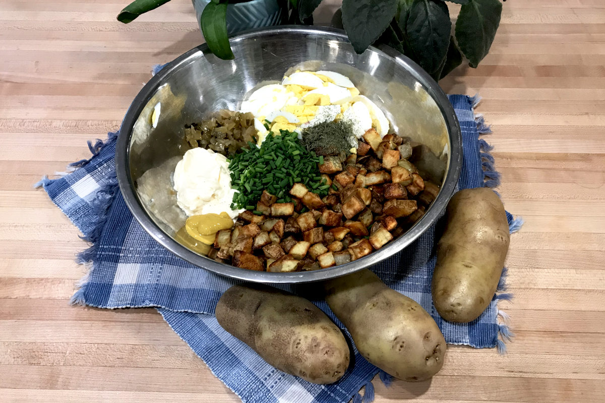 stainless steel bowl with ingredients for homemade potato salad on place mats with whole potatoes and potted plant on butcher block