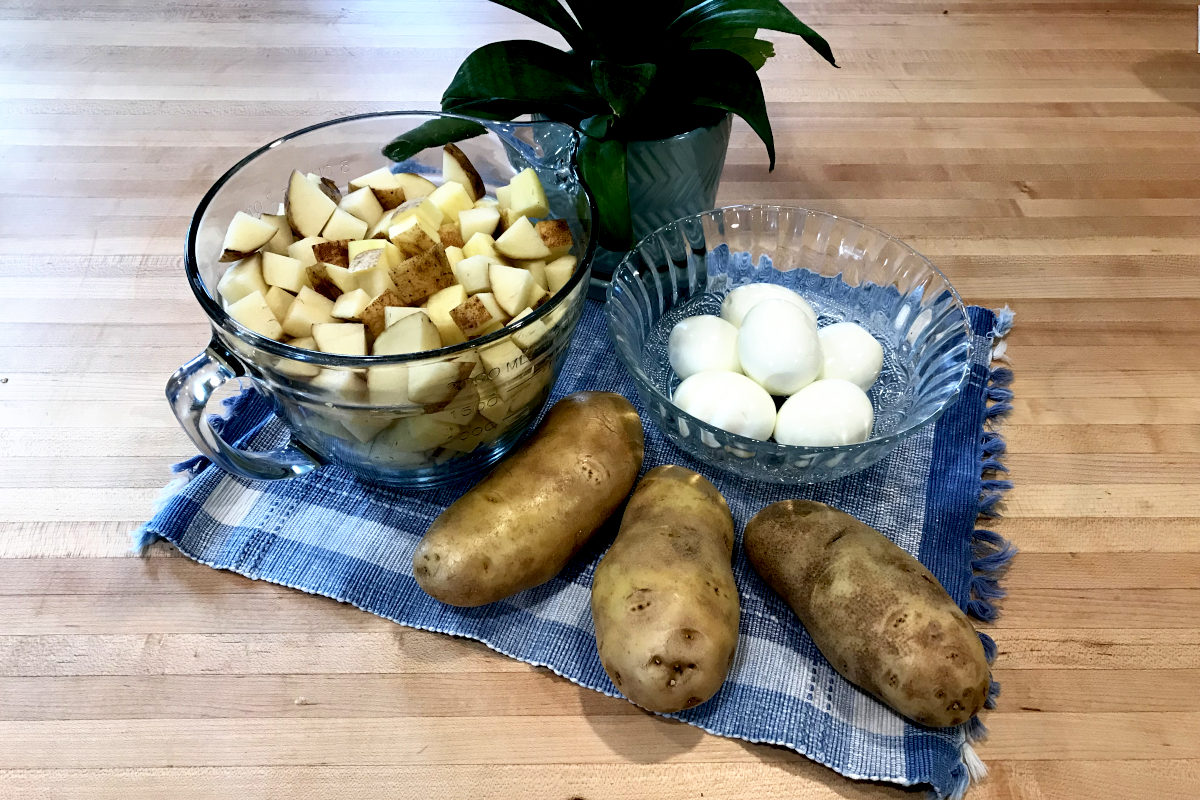 Bowl of diced potatoes, bowl of hard boiled eggs, with whole potatoes and a potted plant on a place mat on butcher block