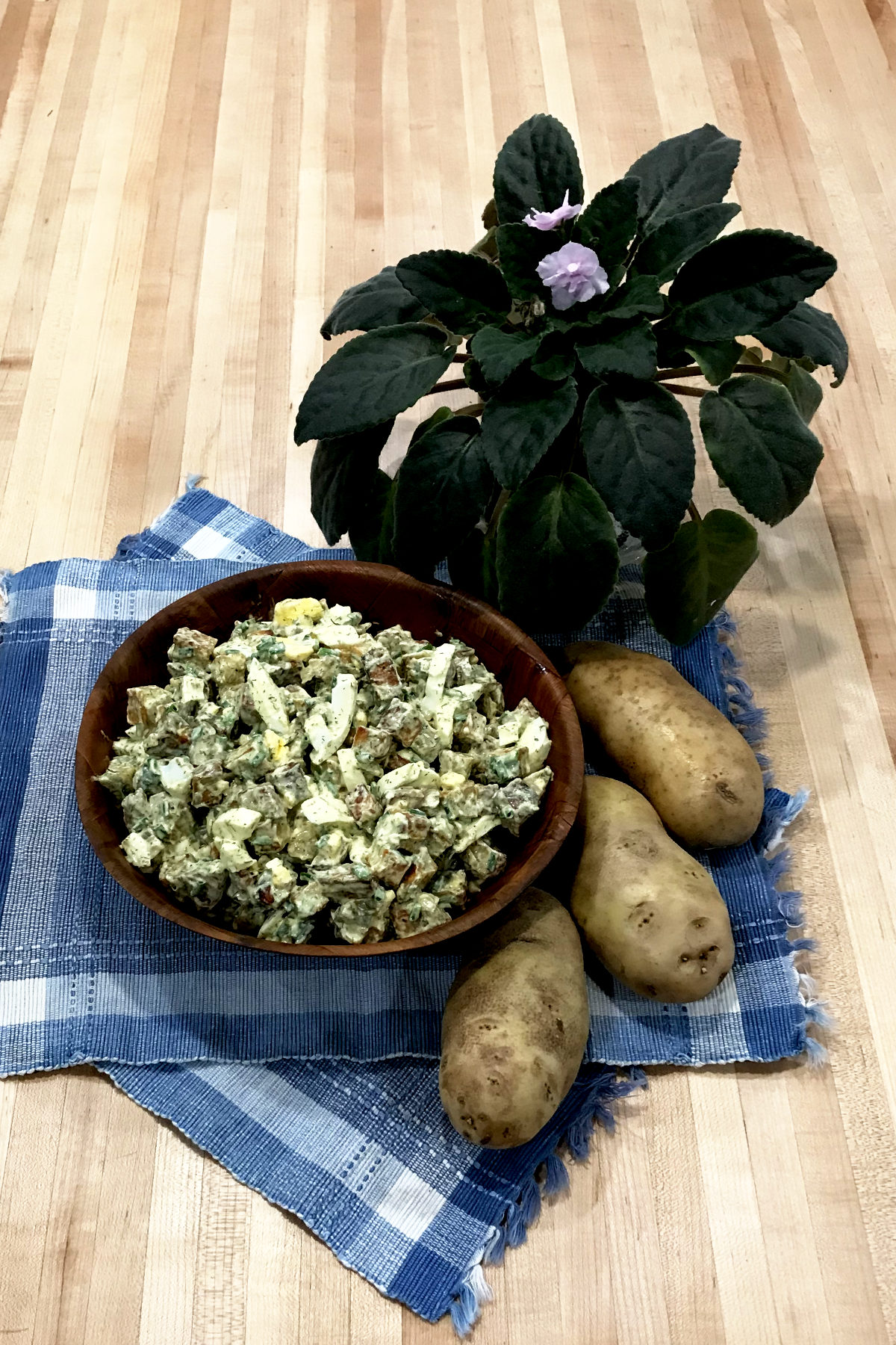 Homemade Potato Salad in wooden bowl on place mat with three potatoes and a potted plant on butcher block