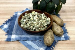 potato salad in wooden bowl on place mats with whole potatoes and potted plant on butcher block