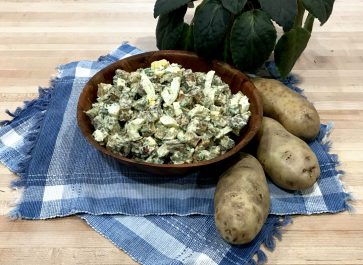 potato salad in wooden bowl on place mats with whole potatoes and potted plant on butcher block