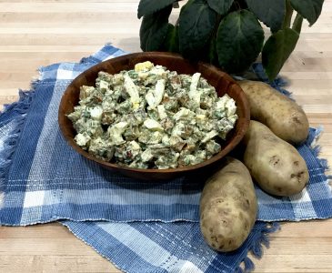potato salad in wooden bowl on place mats with whole potatoes and potted plant on butcher block