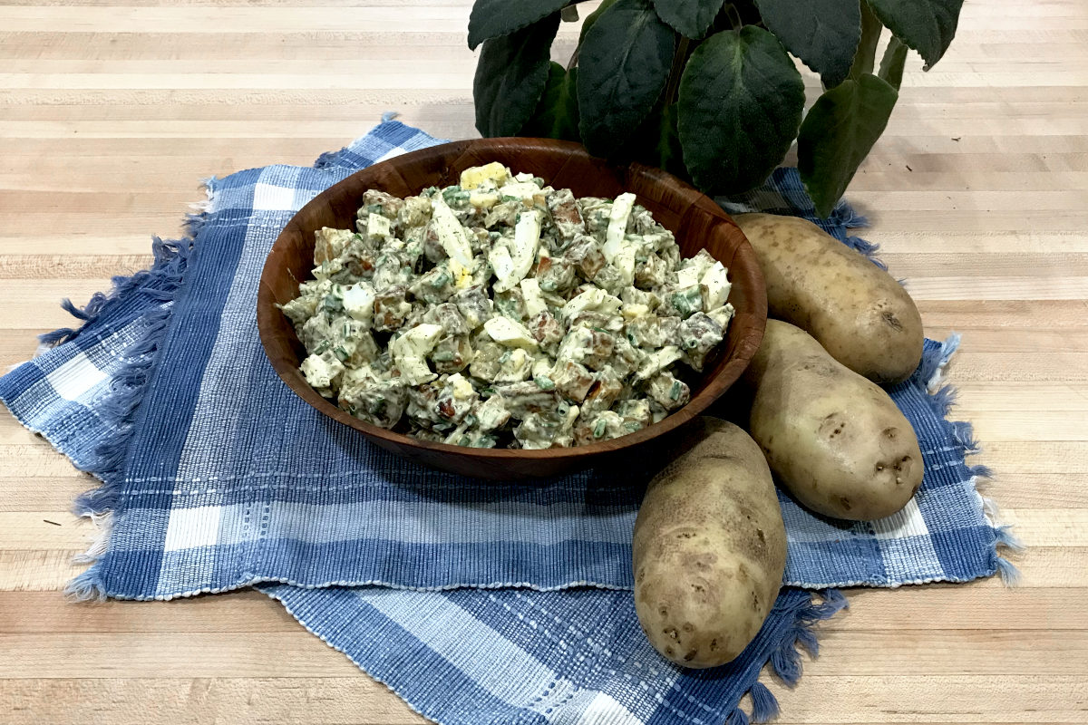 potato salad in wooden bowl on place mats with whole potatoes and potted plant on butcher block.